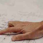 Primo piano su una mano di un uomo con la giacca e camicia che indica un disegno di una linea industriale