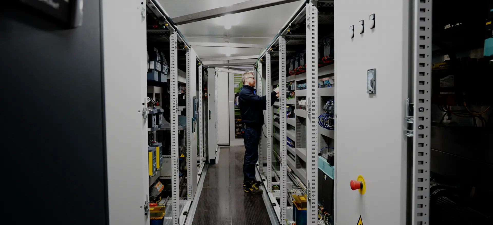 Operatore ispeziona scaffali tecnici in un corridoio di impianto industriale o data center.