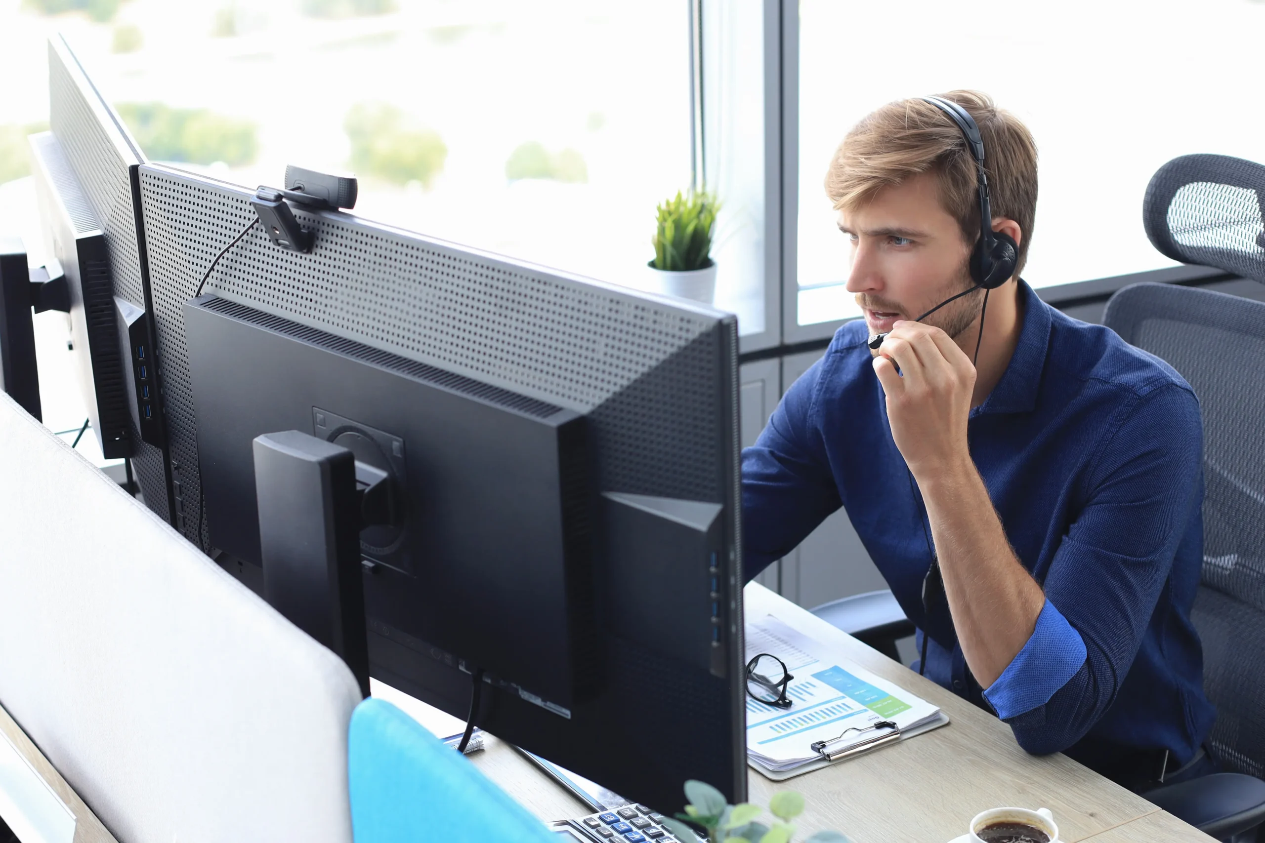 Uomo con cuffie microfoniche lavora al computer in un ufficio moderno con luce naturale.